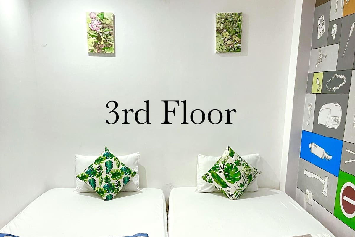 A 3rd floor room with two beds, each with a floral pillow and a checkered blanket. There is a welcome mat with a floral design in front of the beds.