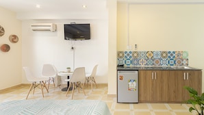 Een compacte hotelkamer met een bed, een eettafeltje met stoelen, een aan de muur bevestigde televisie, een kitchenette met een koelkast, en een betegelde spatwand.