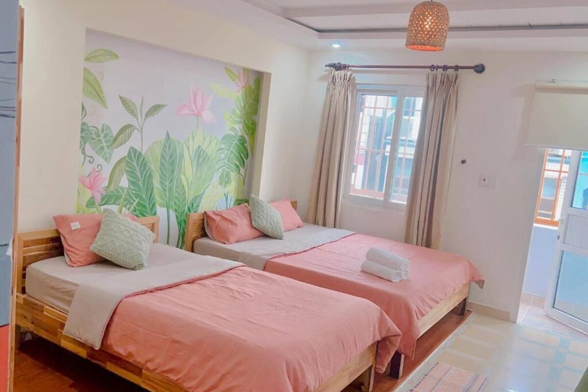 A room with two wooden beds, each with pink bedding and pillows, a large floral mural on the wall, and a window with curtains.