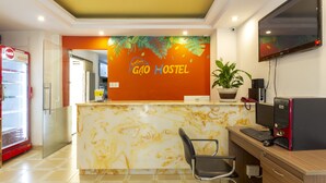 Een receptiebalie met een bord waarop 'Sao Gon Hostel' staat, een computerinrichting, een koelkast en een potplant.