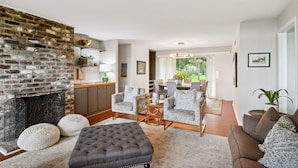 A living room with a brick fireplace, grey upholstered chairs, a tufted ottoman, and a dining area with a table and chairs. - Waterfront “State of Mind” vintage home.  (Bluffton)