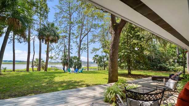 A wooden deck with outdoor furniture, overlooking a grassy area with trees and a view of the water. - Waterfront “State of Mind” vintage home.  (Bluffton)