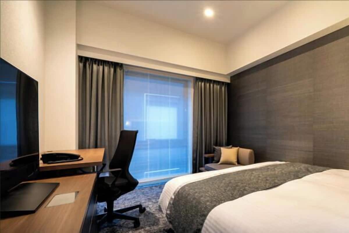 Een hotelkamer met een bed, een bureau, een stoel, een televisie en een groot raam met gordijnen.