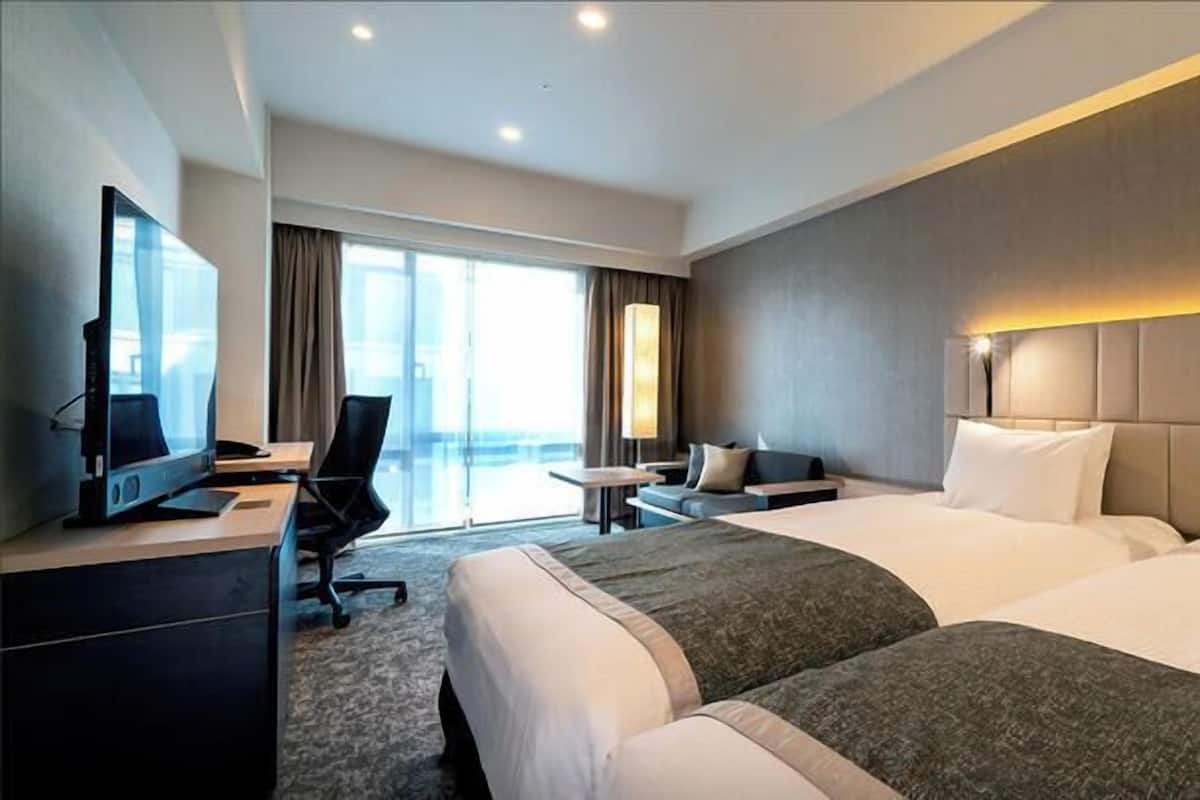 Een hotelkamer met twee bedden, een bureau met een computer, een televisie en een groot raam met gordijnen.