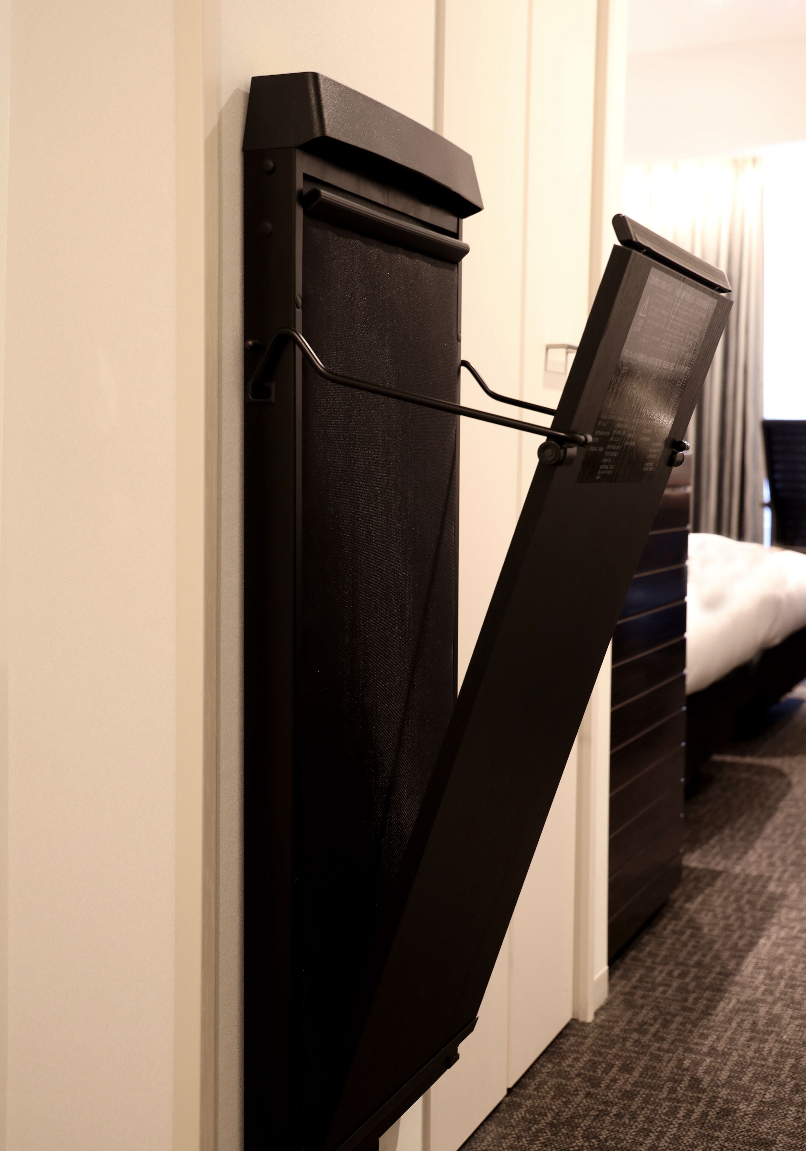 Un bureau compact, pliable et fixé au mur, doté d’une surface plane et d’une poignée, placé dans un couloir étroit, avec un sol recouvert d’une moquette et un lit en arrière-plan.