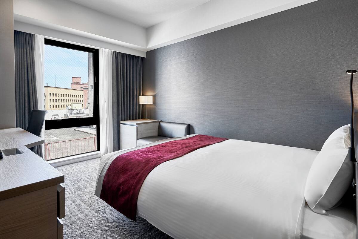 Una habitación de hotel moderna con una cama grande, un escritorio y una silla. Hay una ventana con cortinas y vistas a edificios.