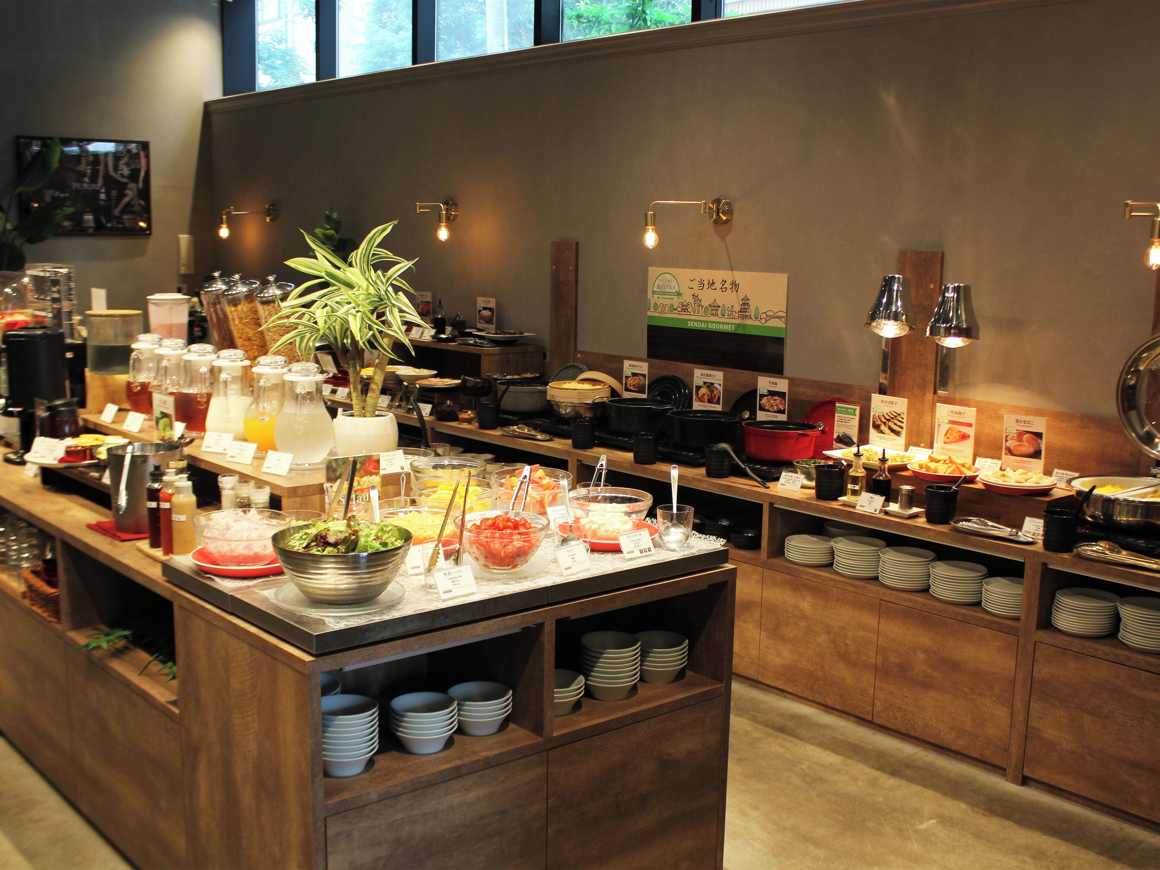 A buffet setup with various food items and beverages, including a plant centerpiece, in a well-lit room with wooden furniture and a sign indicating a local delicacy.