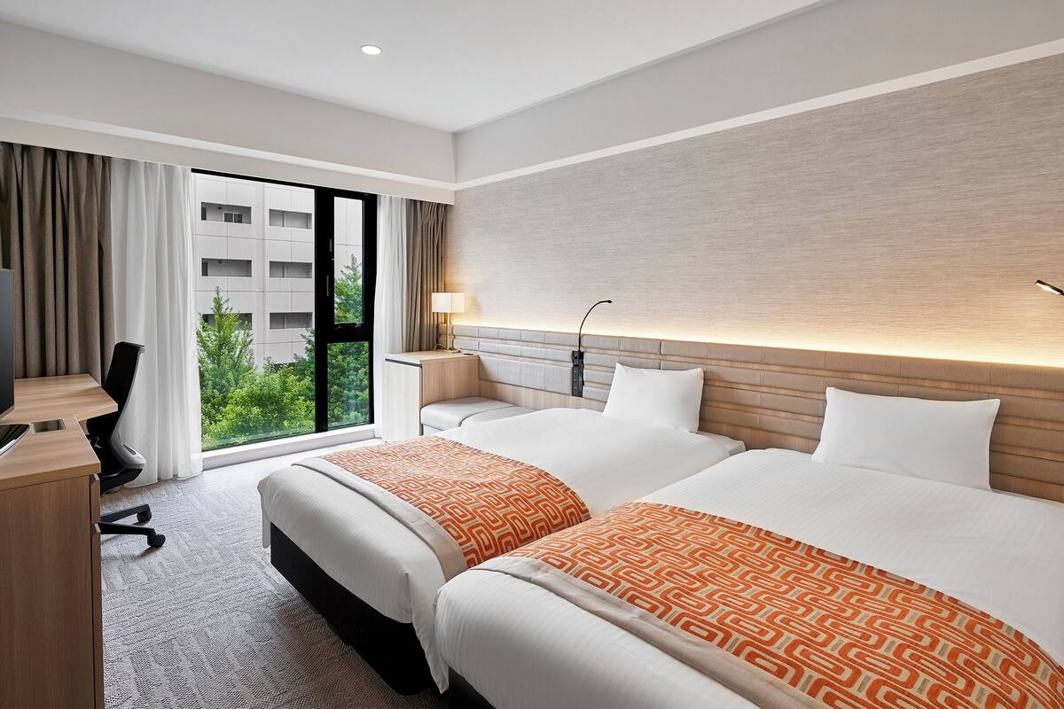 Habitación de hotel con dos camas, un escritorio, una silla, una lámpara y vista a zonas verdes.