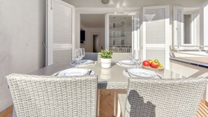 Un espace extérieur aménagé pour les repas, avec des chaises en osier et une table en verre, offrant une vue sur l’immeuble voisin et agrémenté d’une plante en pot posée sur la table.