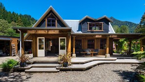 A wooden lodge with a covered porch, a dining area, and a view of a forested mountain. - On The Track Lodge (Nydia Bay)