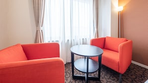 Een hotelkamer met twee oranje fauteuils, een klein rond tafeltje, een groot raam met dunne gordijnen en een vloerlamp.