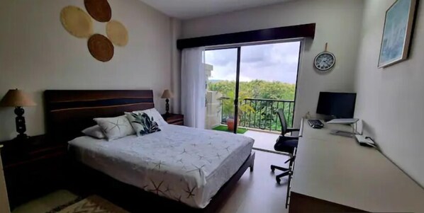 A bedroom with a large bed, a desk, a chair, a TV, and a balcony with a view of greenery.