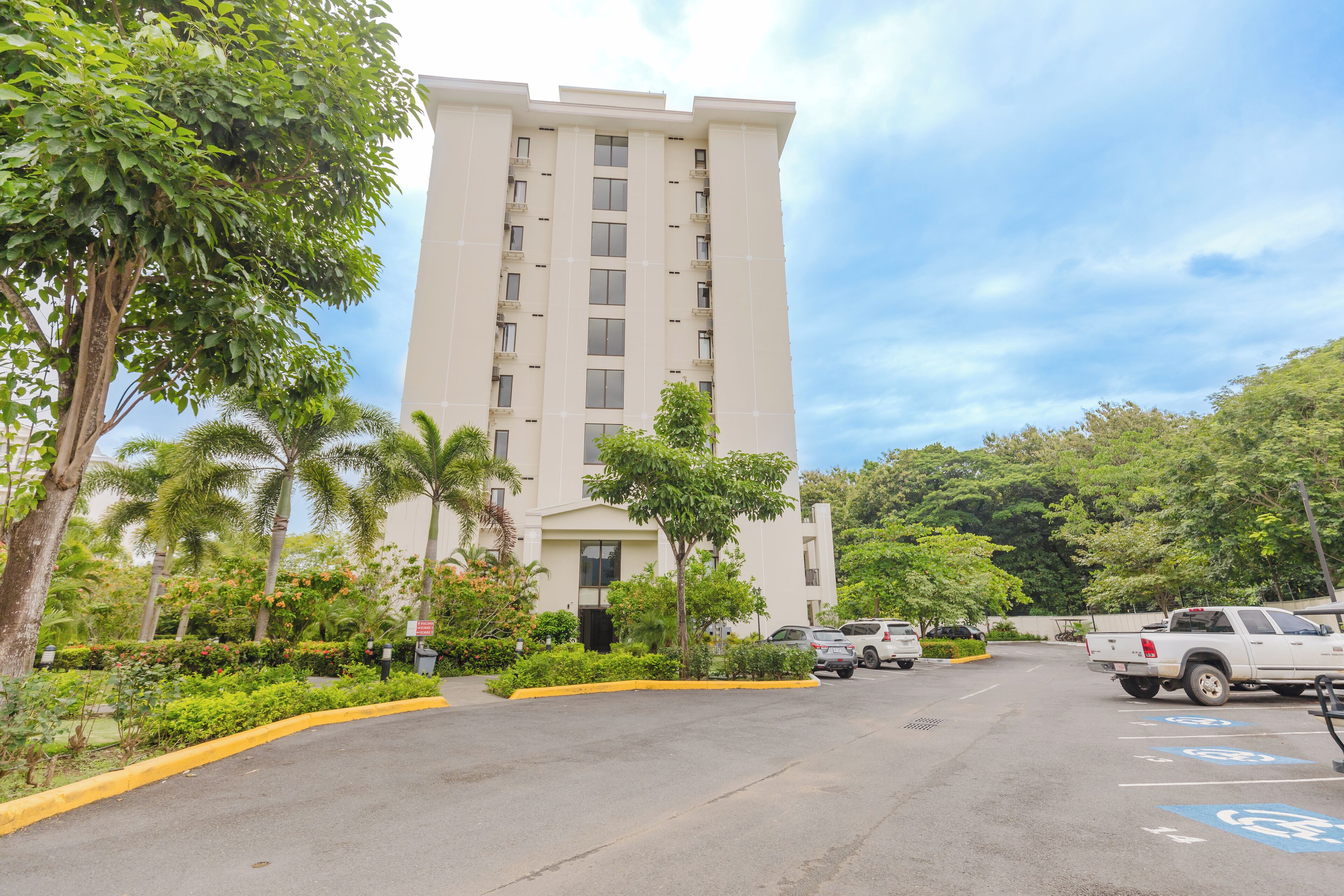 A parking lot with a tall building in the background, surrounded by greenery and palm trees.