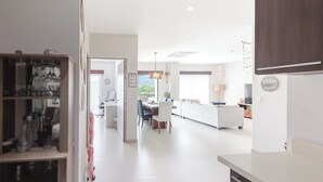 A modern kitchen with white countertops, a dining area with a white table and chairs, and a view of the outdoors through a large window. - Lovely Mountain View Apartment (Jacó)