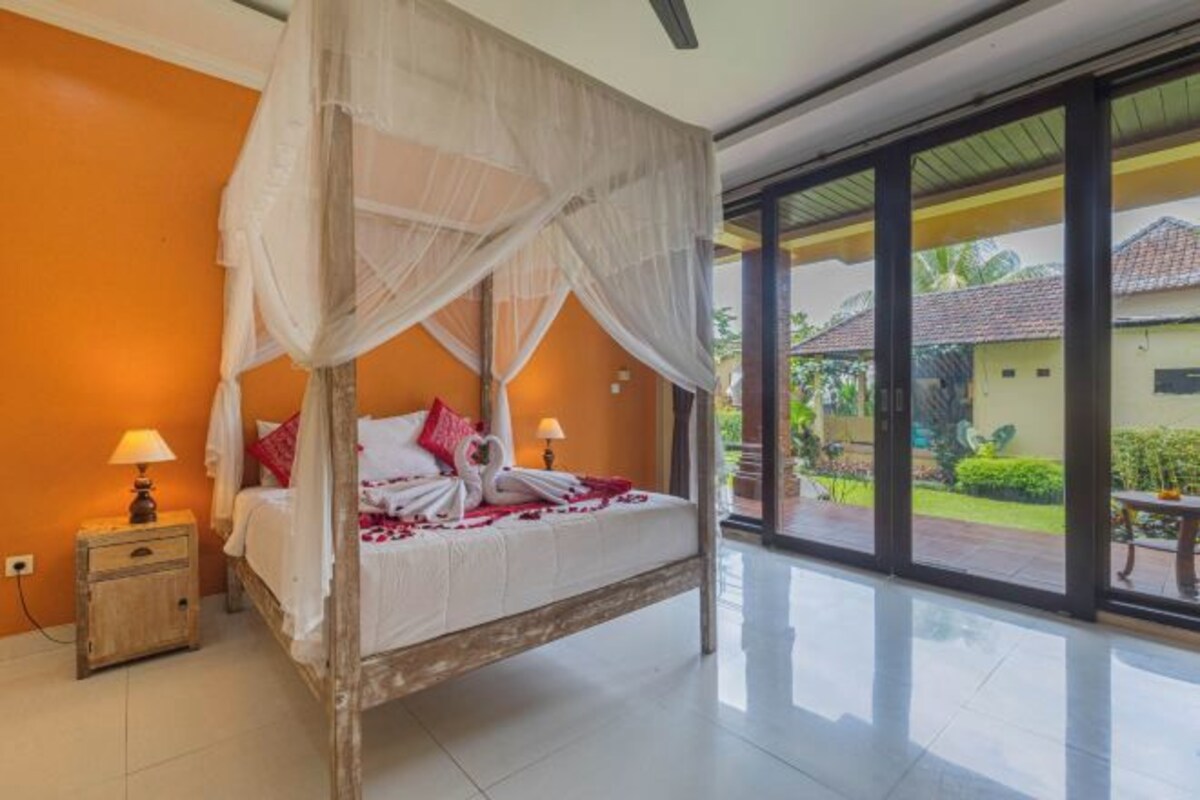 A four-poster bed with white bedding and red pillows, a nightstand with a lamp, and a view of a garden through large glass doors.