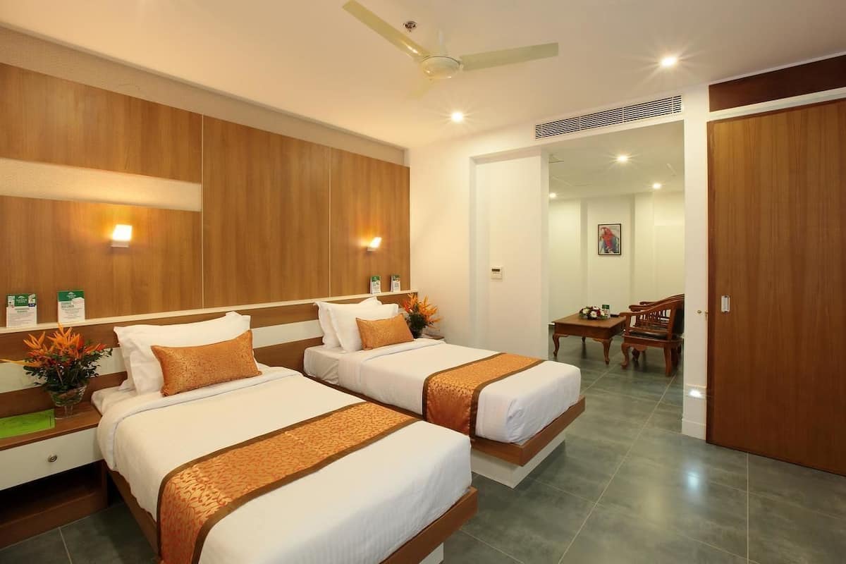 Quarto de hotel com duas camas, um ventilador de teto, área de refeições com mesa e cadeiras, e uma porta de madeira.