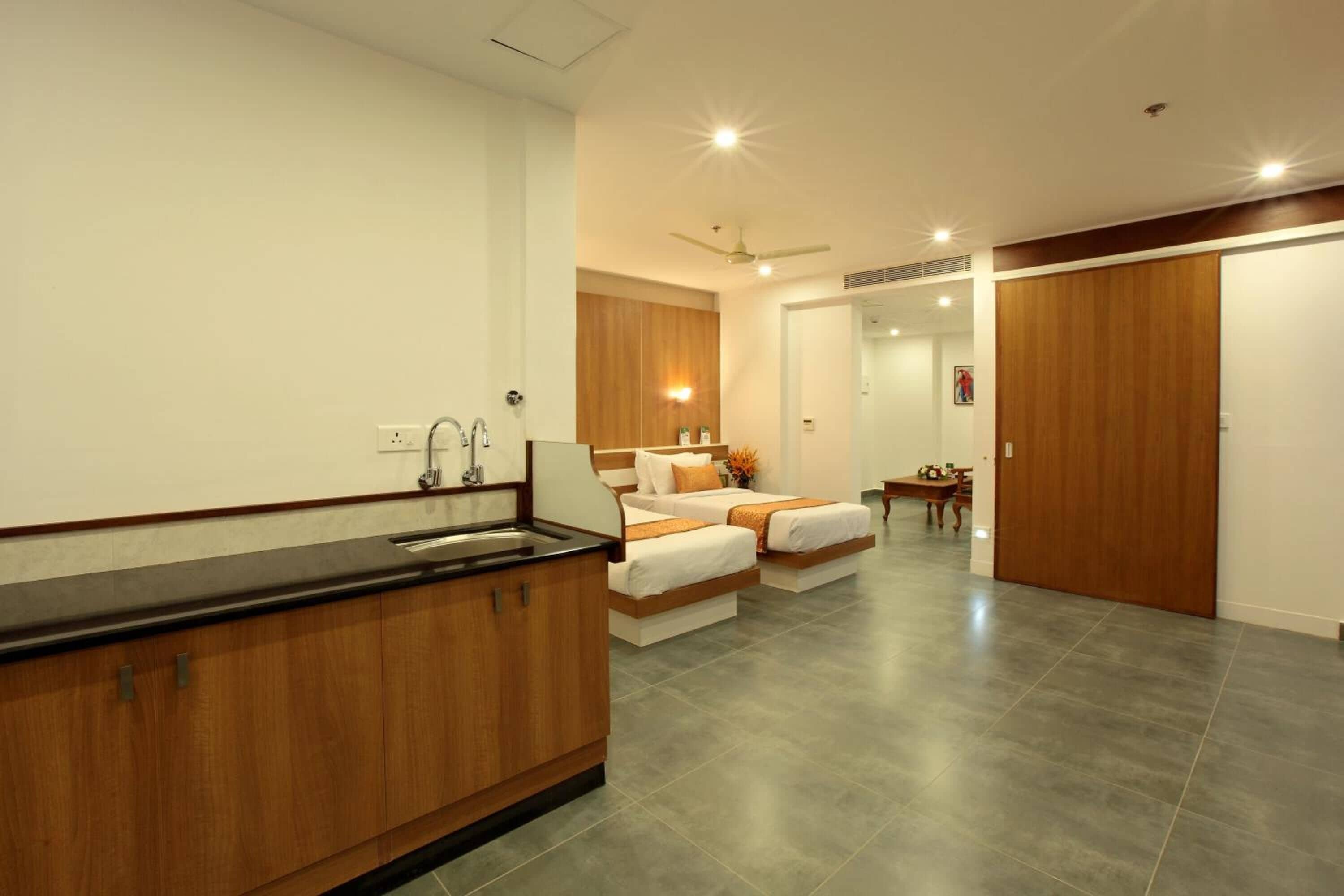 A modern hotel room with a kitchenette, two beds with orange bedding, a wooden wardrobe, and a dining area.
