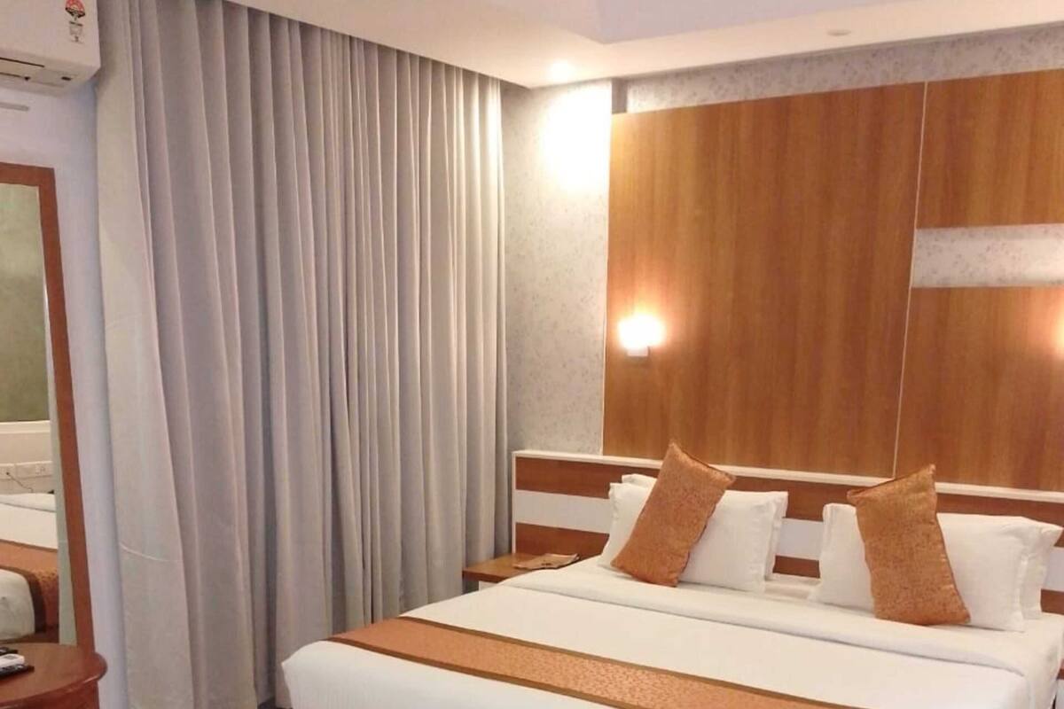 Quarto de hotel com uma cama grande, cabeceira de madeira, mesa de cabeceira e ar condicionado fixado na parede.