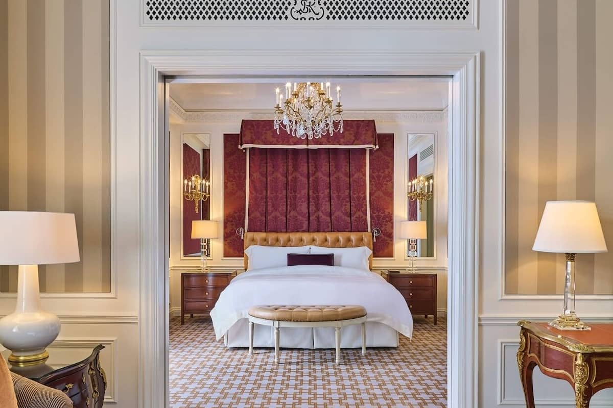 A hotel room with a large bed, a chandelier, a red headboard, and a patterned floor.