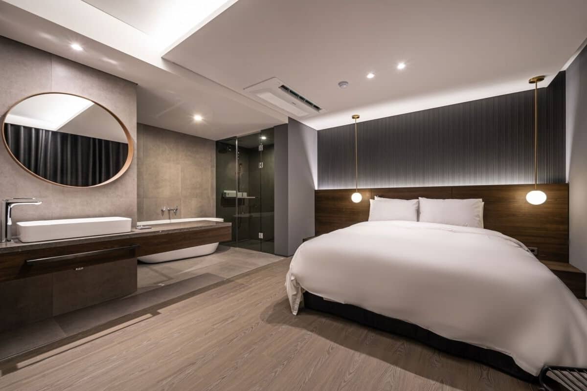 A modern hotel room with a large bed, a round mirror, a vanity with a sink, and a shower area.