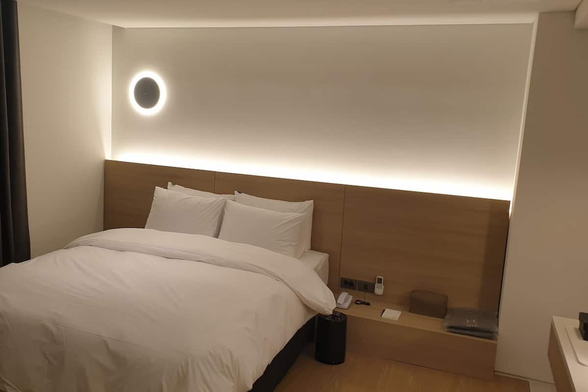 A modern hotel room with a neatly made bed, a round wall light, a bedside table with a phone, and a trash bin.
