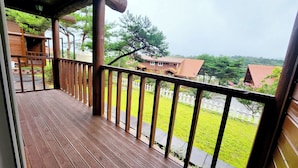 A wooden balcony with a view of a white picket fence, green grass, and houses with red roofs.