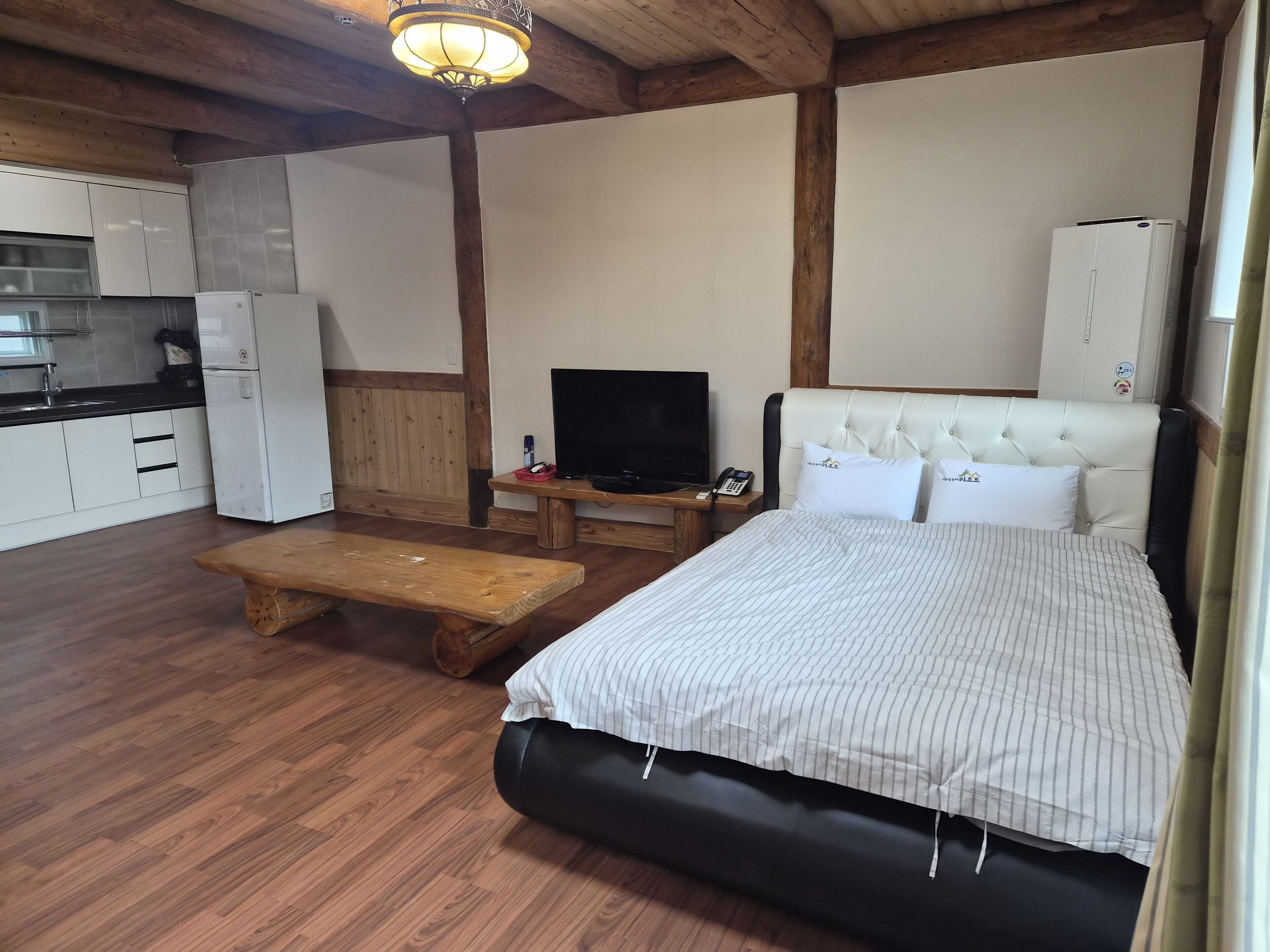 A room with a bed, a television, a wooden bench, a small table, and a kitchenette.