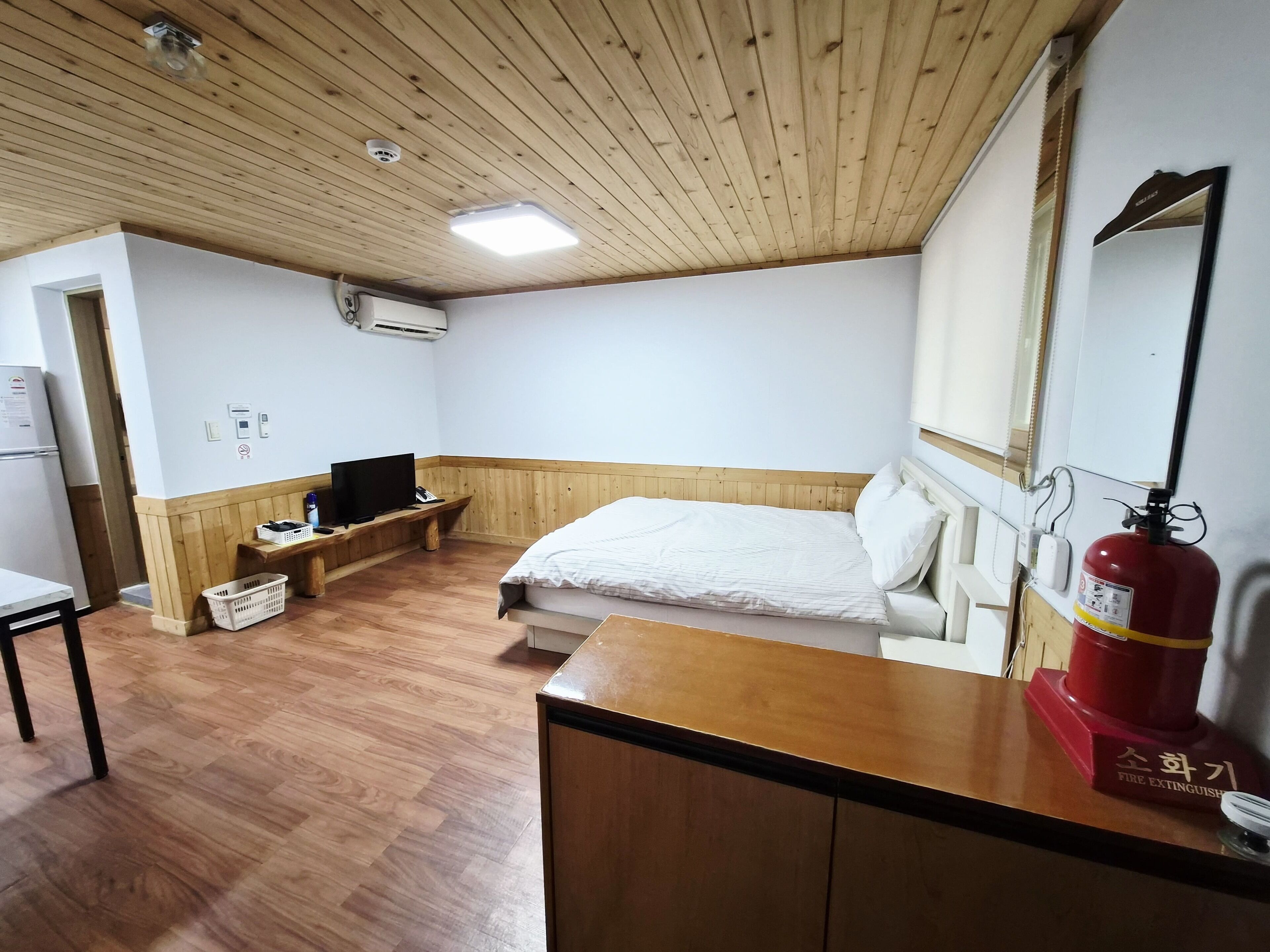 Quarto com teto de madeira, cama com roupa de cama branca, mesa de cabeceira de madeira, televisão, extintor de incêndio e parede branca com janela.