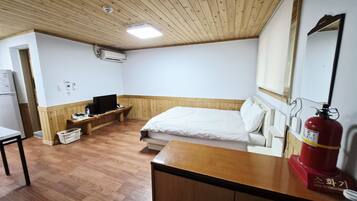 Quarto com teto de madeira, cama com roupa de cama branca, mesa de cabeceira de madeira, televisão, extintor de incêndio e parede branca com janela.