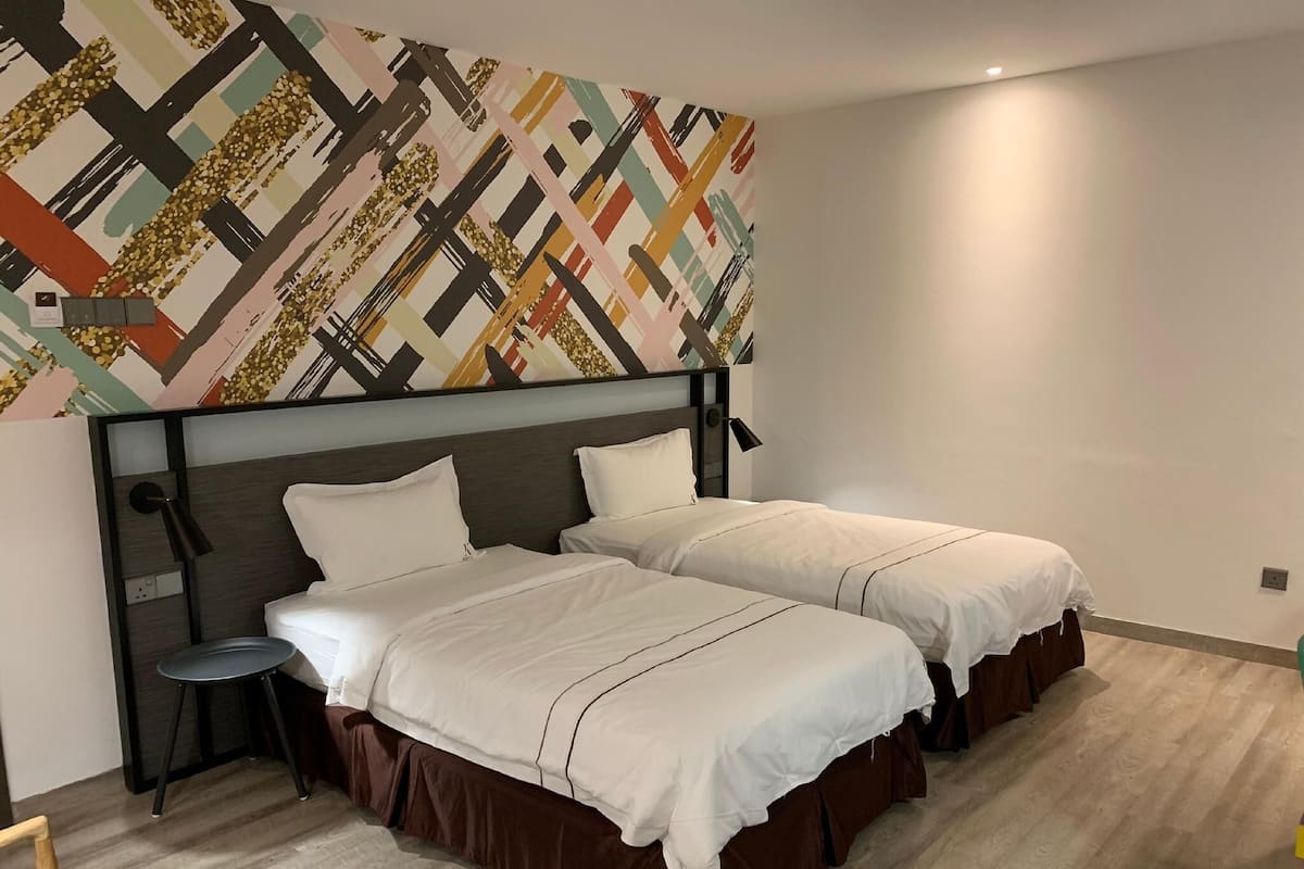 A hotel room with two beds, a patterned wall, a small stool, and a wooden floor.