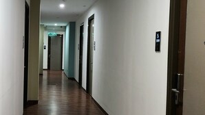 Un pasillo largo y estrecho con suelo de madera y paredes blancas. Hay puertas a ambos lados, algunas con letreros encima.