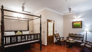 Uma cama com dossel, estrutura de madeira, e um banquinho no canto. Há uma porta com espelho e um quadro na parede.