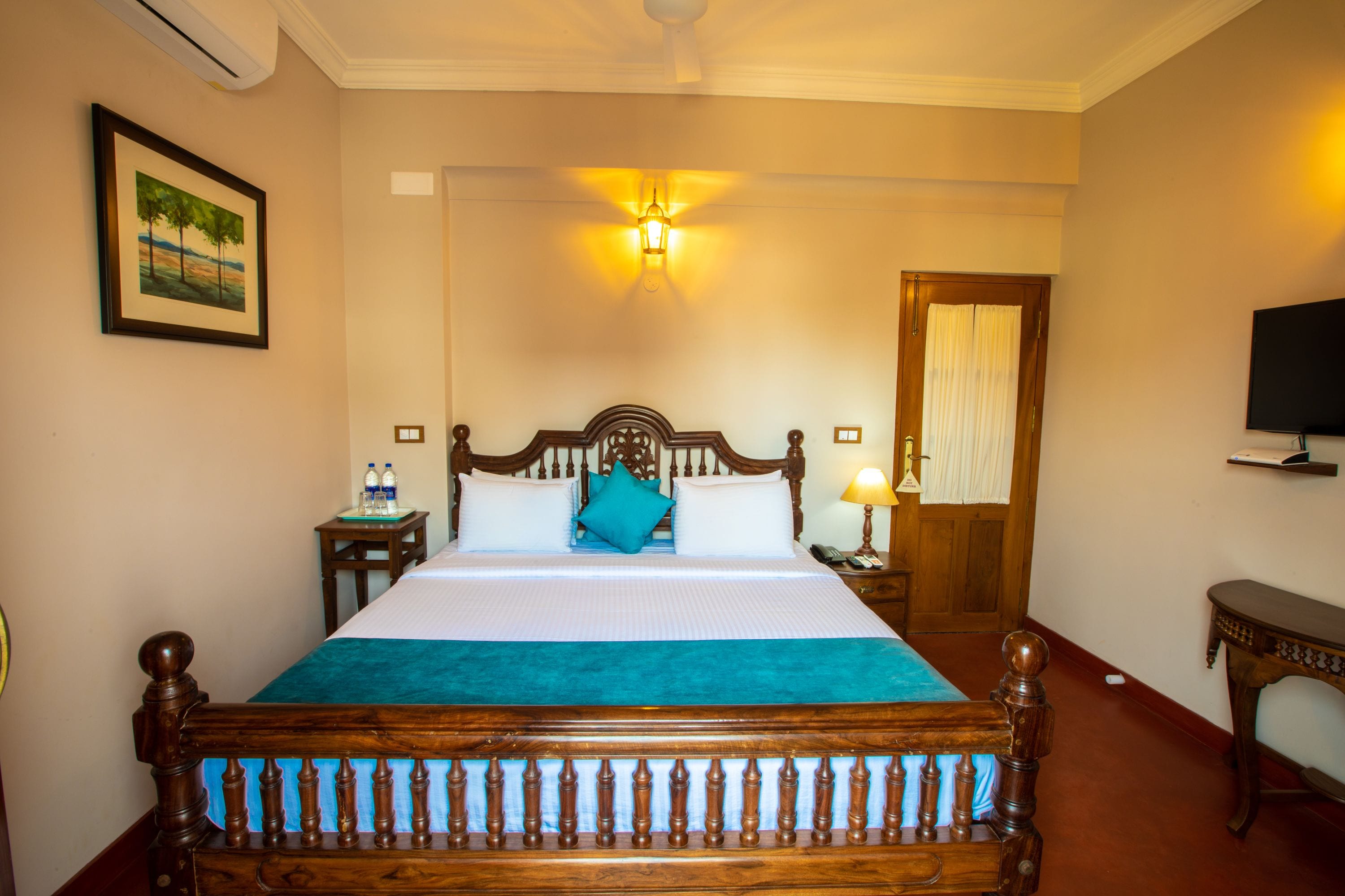 A hotel room with a wooden bed, a bedside table with a lamp, a framed picture on the wall, and a television on the wall.