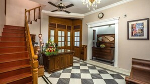 Uma escadaria com corrimãos de madeira, um ventilador de teto, um lustre, uma mesa de recepção com um arranjo floral, um chão quadriculado, uma cômoda de madeira e um quadro na parede.