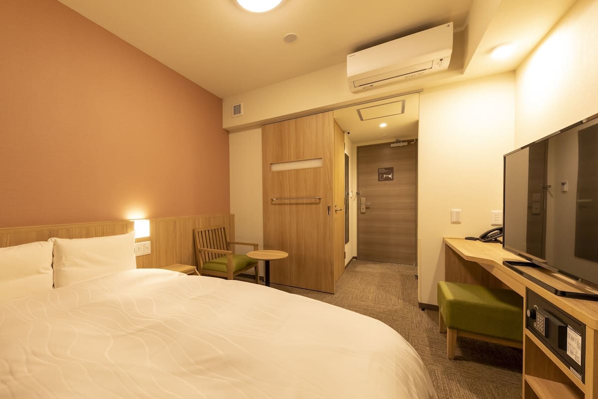 A hotel room with a large bed, a desk with a television, a chair, and an air conditioning unit.