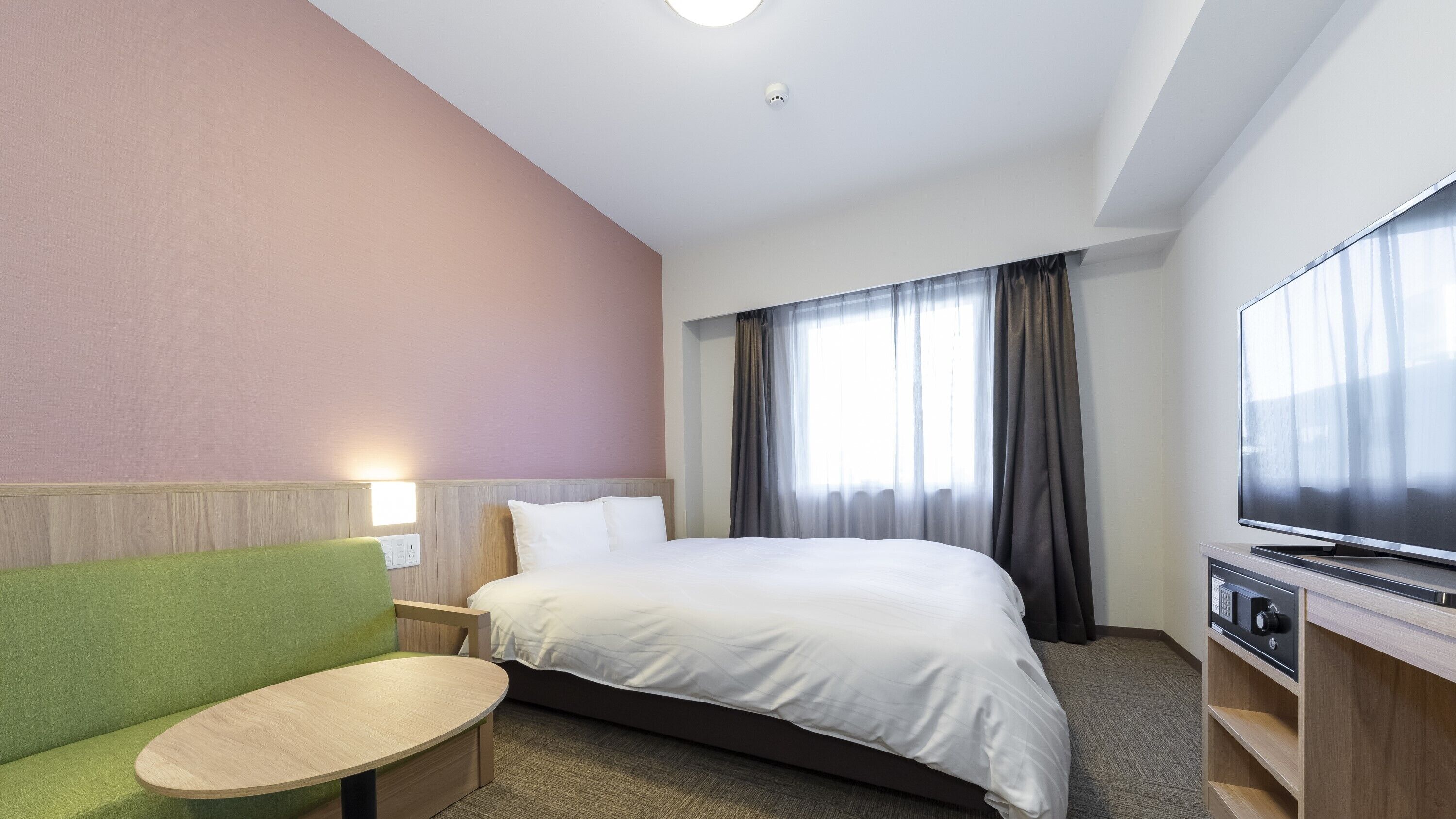 A hotel room with a bed, a TV, a green sofa, a small table, and a window with curtains.