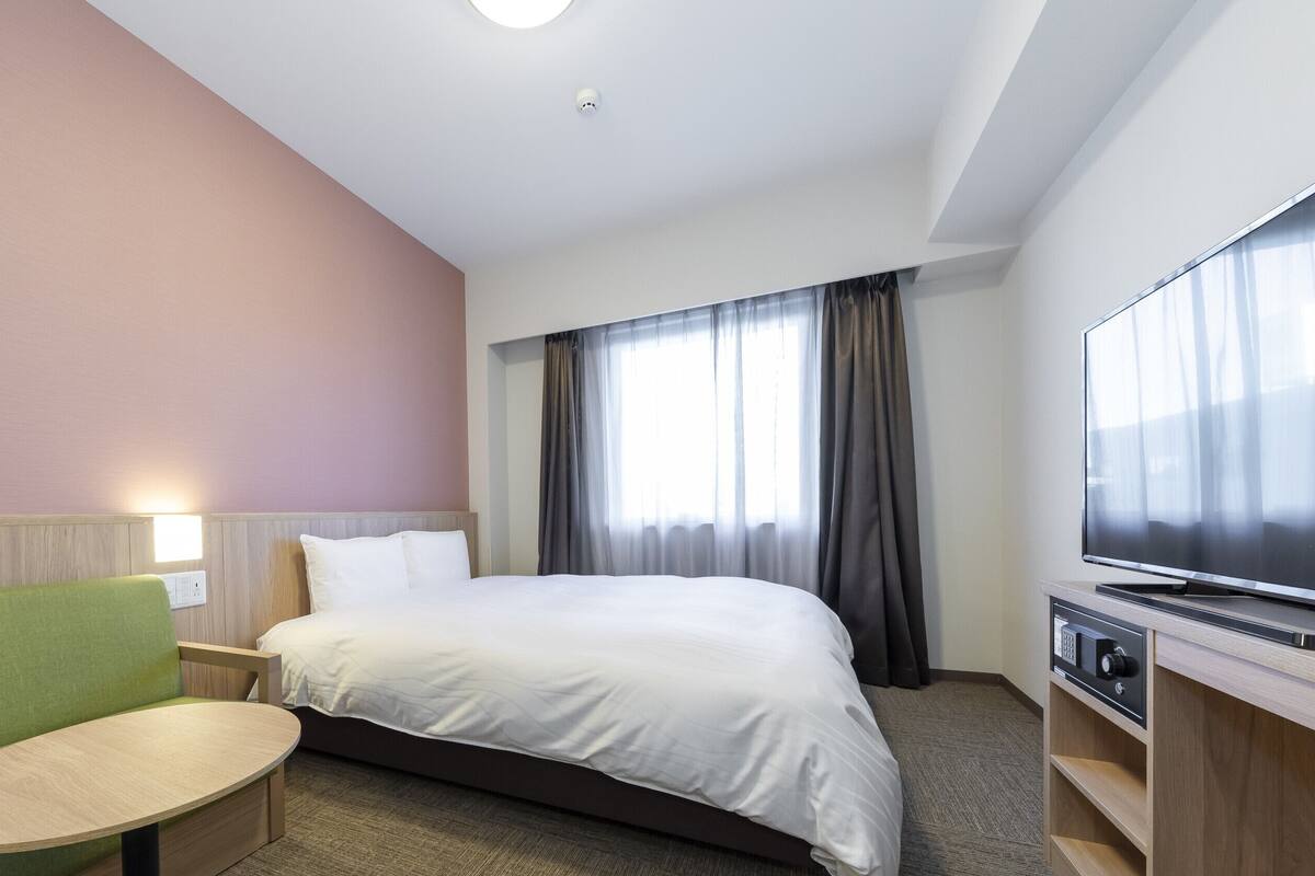 A hotel room with a bed, a TV, a green sofa, a small table, and a window with curtains.