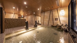 A spa area with a wooden ceiling, stone walls, a hot tub, and a view of the city through a glass door. - Dormy Inn mito Hot Springs (Mito)