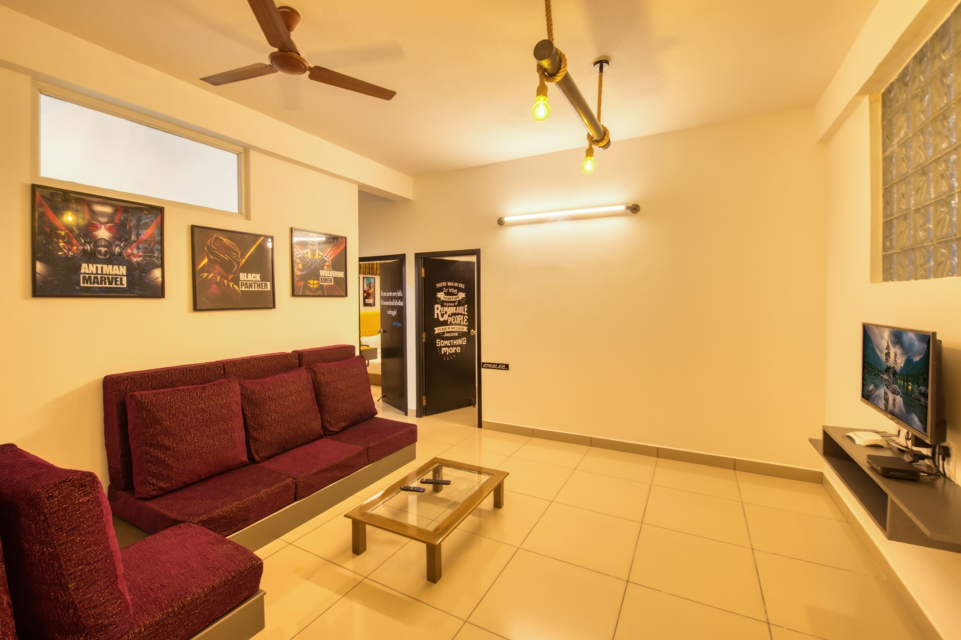 A living room with a red sofa, a coffee table, and a TV on a stand. There are posters on the wall and a ceiling fan.