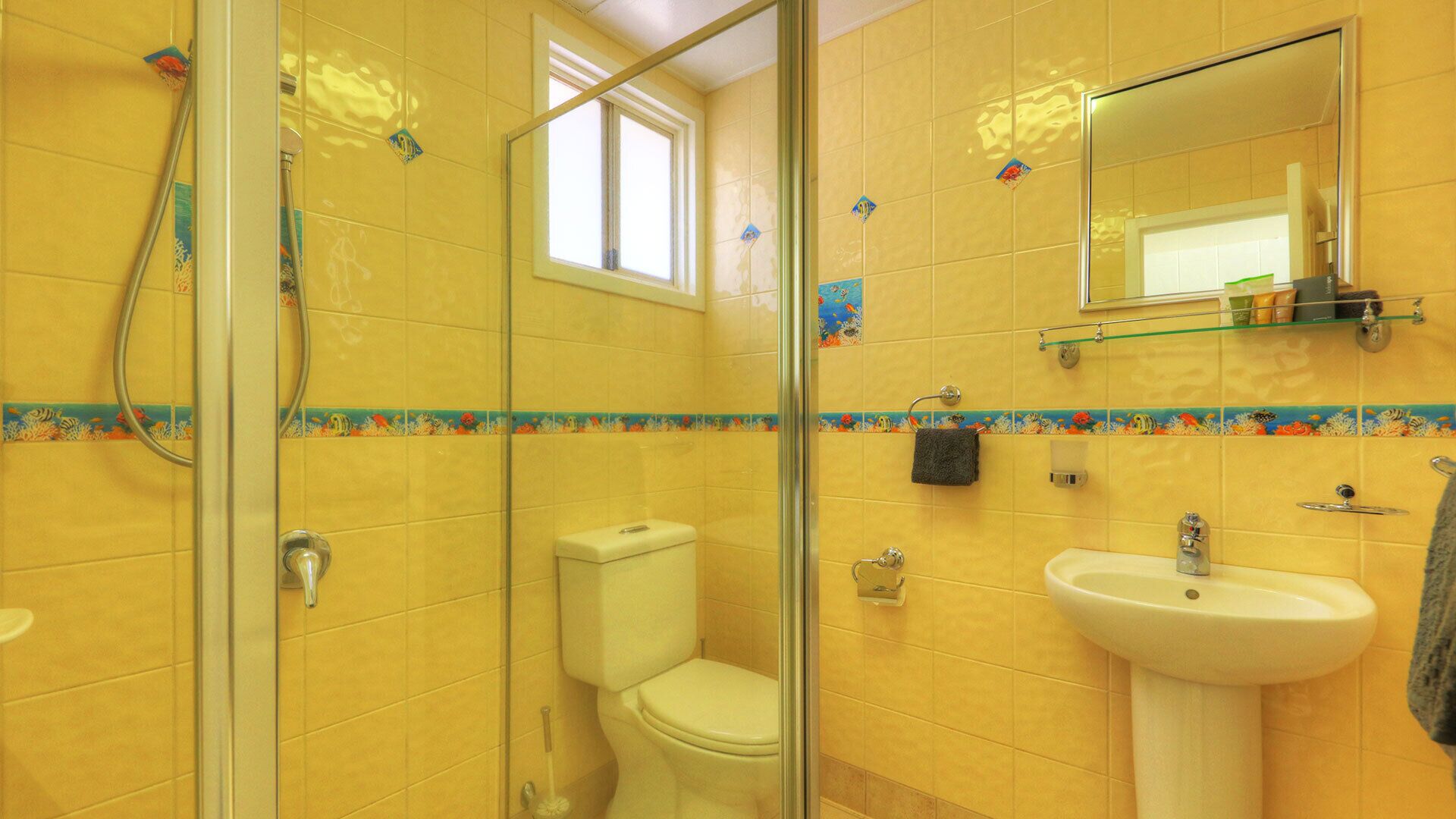 Ein Badezimmer mit gelben Fliesen, einer Duschkabine aus Glas, einer Toilette, einem Waschbecken und einem Spiegel.