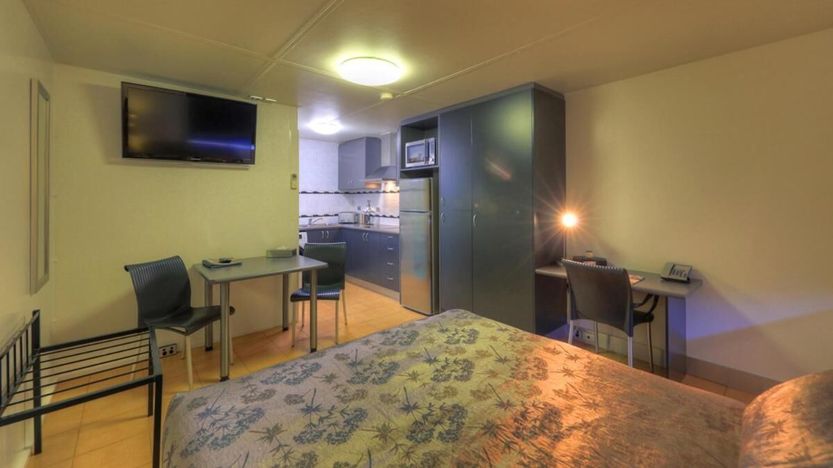 Una habitación pequeña con cama, escritorio, silla, televisor y kitchenette.