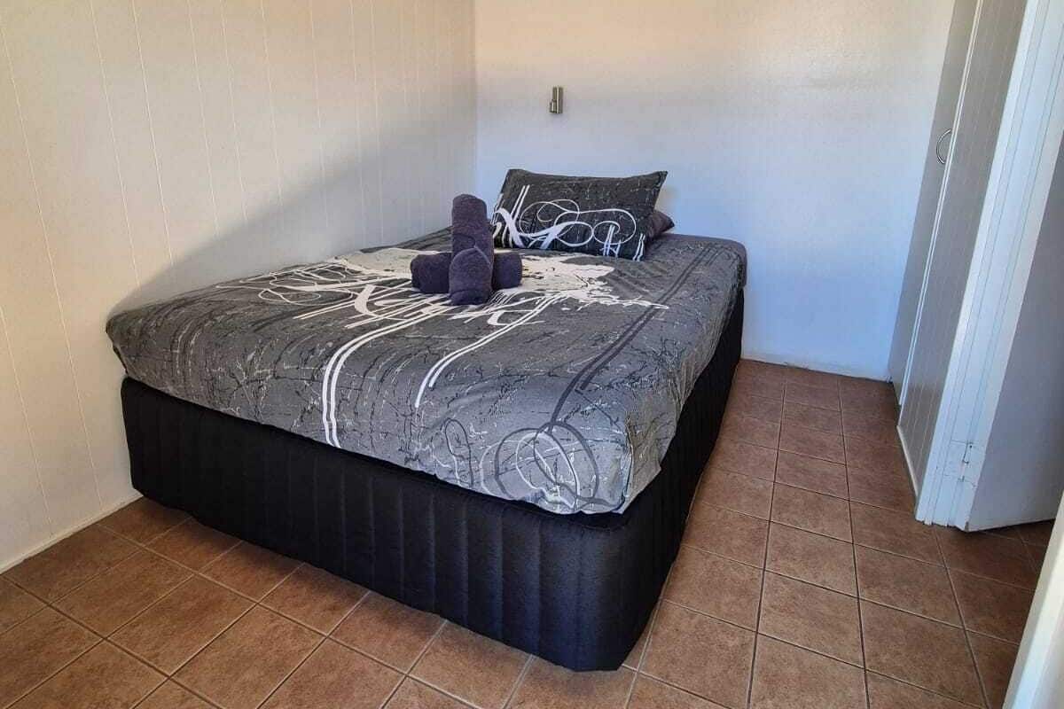 A small, single-bed room with a black mattress and a patterned blanket. There is a wall-mounted air conditioning unit above the bed.