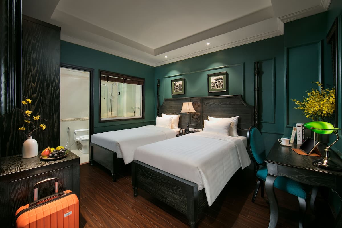 Une chambre d’hôtel avec deux lits, un bureau, une chaise, une petite table avec un vase de fleurs et une valise.