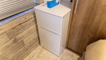 Un refrigerador blanco compacto con una caja azul encima, ubicado en una pequeña cocina con piso y gabinete de madera.