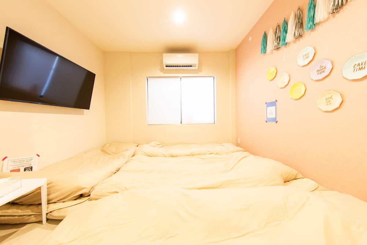 Una habitación con una cama, un televisor de pantalla plana montado en la pared, un aire acondicionado, una ventana y adornos en las paredes, incluyendo carteles con frases como 'cake time' y 'go to sleep'.