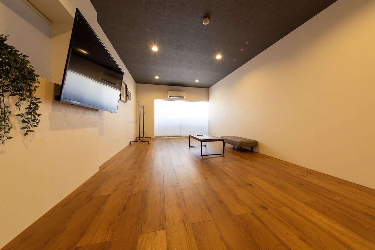 Una habitación amplia con suelo de madera, un televisor de pantalla plana montado en la pared, una mesita y un banco.