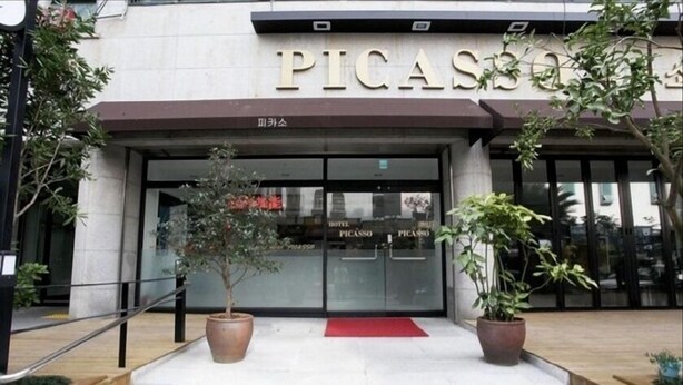 The exterior of a hotel named 'PICASSO' with a modern facade, glass doors, and potted plants in front.
