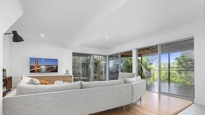 A modern living room with a large sectional sofa, a flat-screen TV, and a large sliding glass door leading to a balcony with a view of greenery. - Stunning Seaviews, Sunshine Beach (Sunshine Beach)