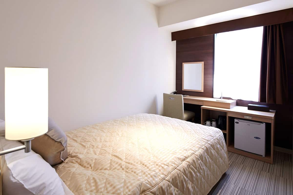 Ein Hotelzimmer mit Bett, Schreibtisch, Stuhl, kleinem Kühlschrank, Lampe und einem Fenster mit Vorhängen.