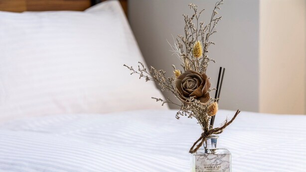 Una cama con un florero de vidrio transparente que contiene flores secas y una rosa marrón.